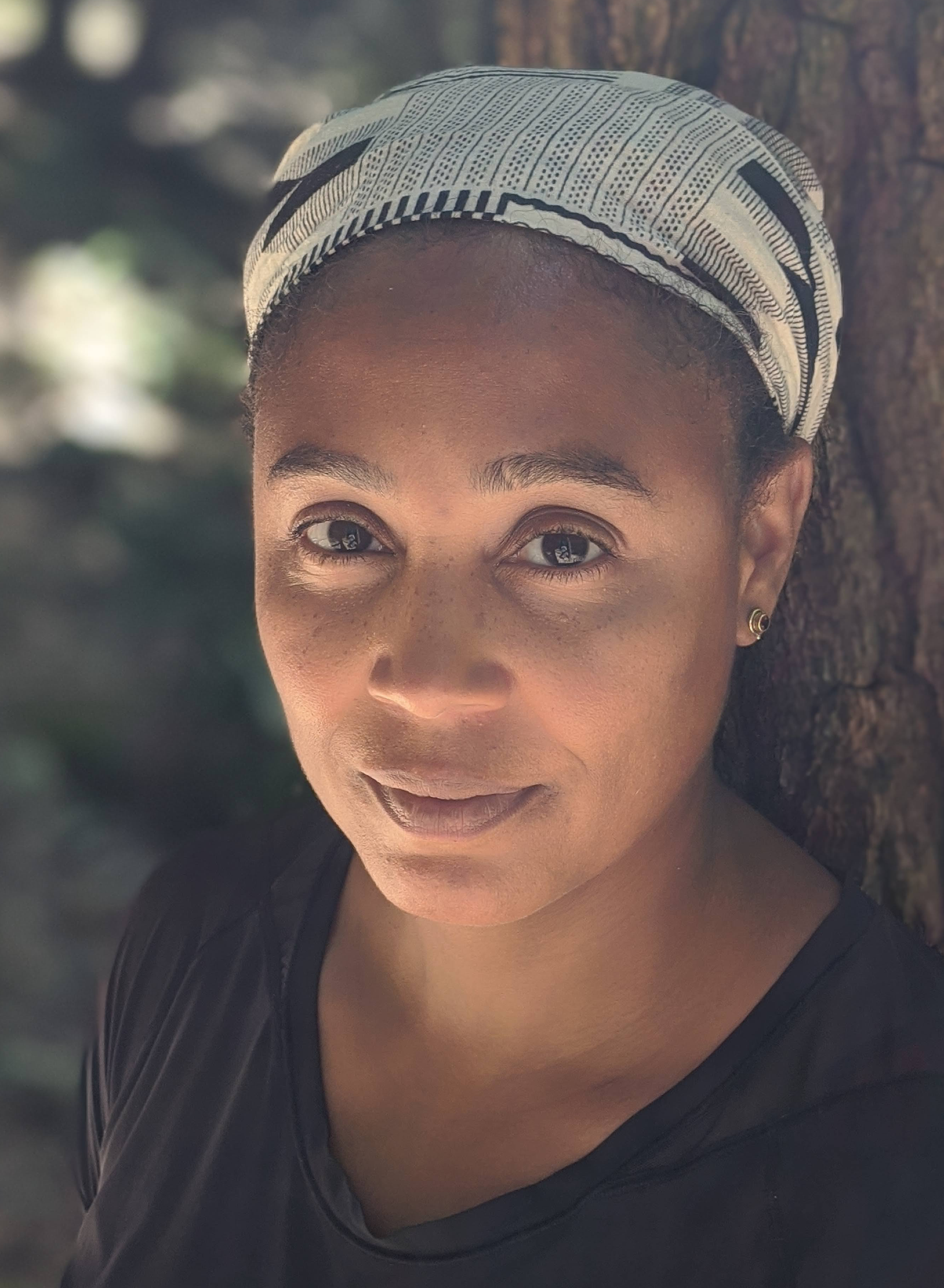 A woman wearing a patterned headscarf and black top stands outdoors leaning against a tree, looking directly at the camera.