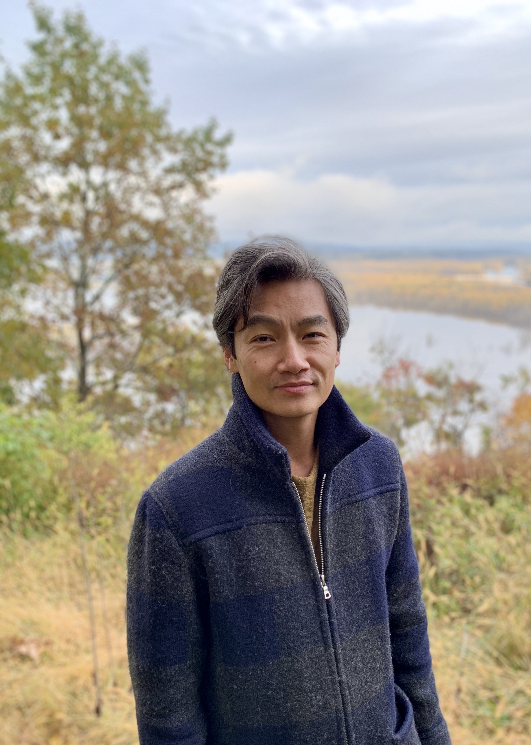 A person standing outdoors with a neutral expression, wearing a dark jacket. Behind them, a scenic view of a lake surrounded by autumn foliage and trees under a cloudy sky.