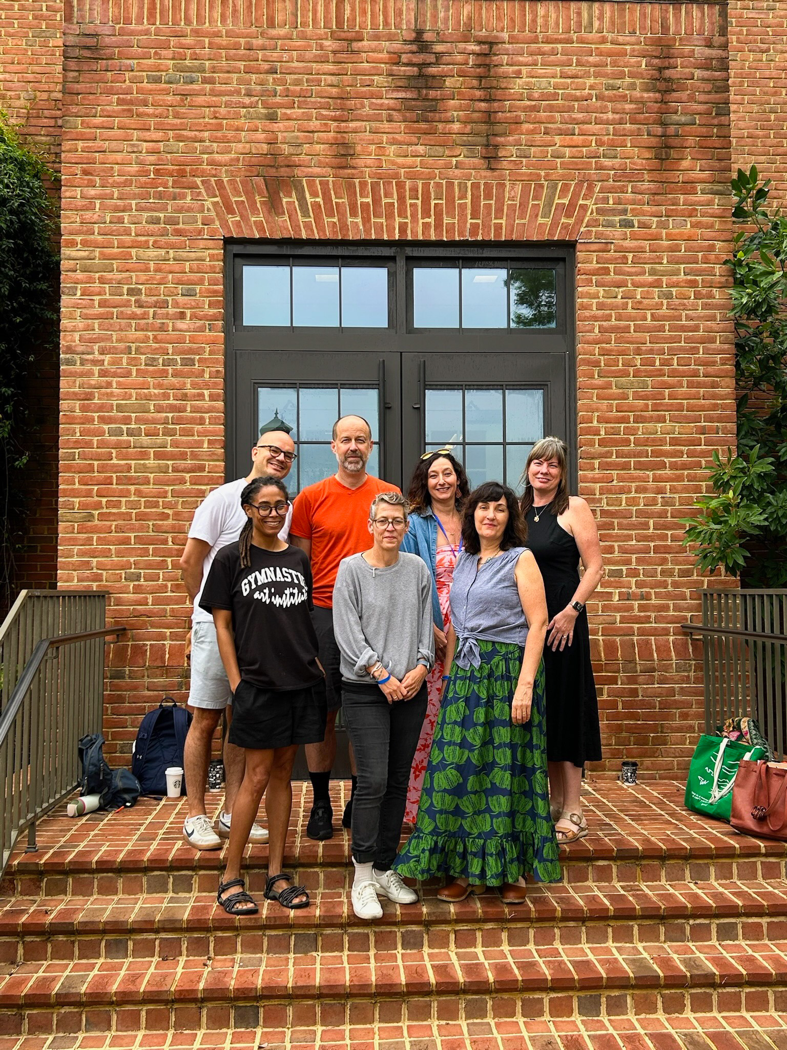 Seven people stand together on brick steps in front of a large windowed door, posing for a group photo. Bags and greenery are visible at the sides.