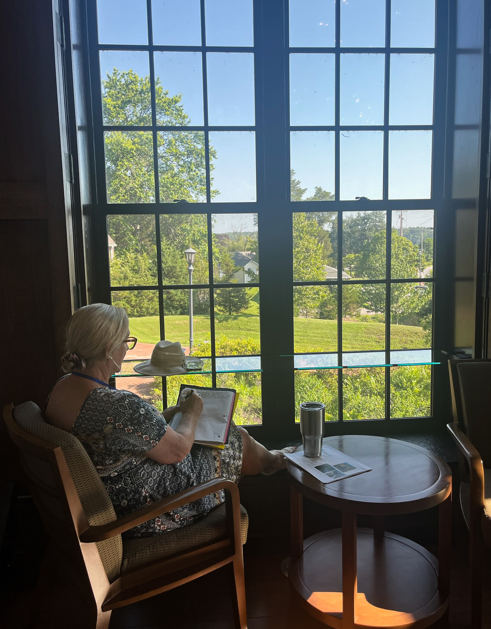 A woman sits by a large window, writing on a clipboard, with sunlight streaming in and a green landscape visible outside.