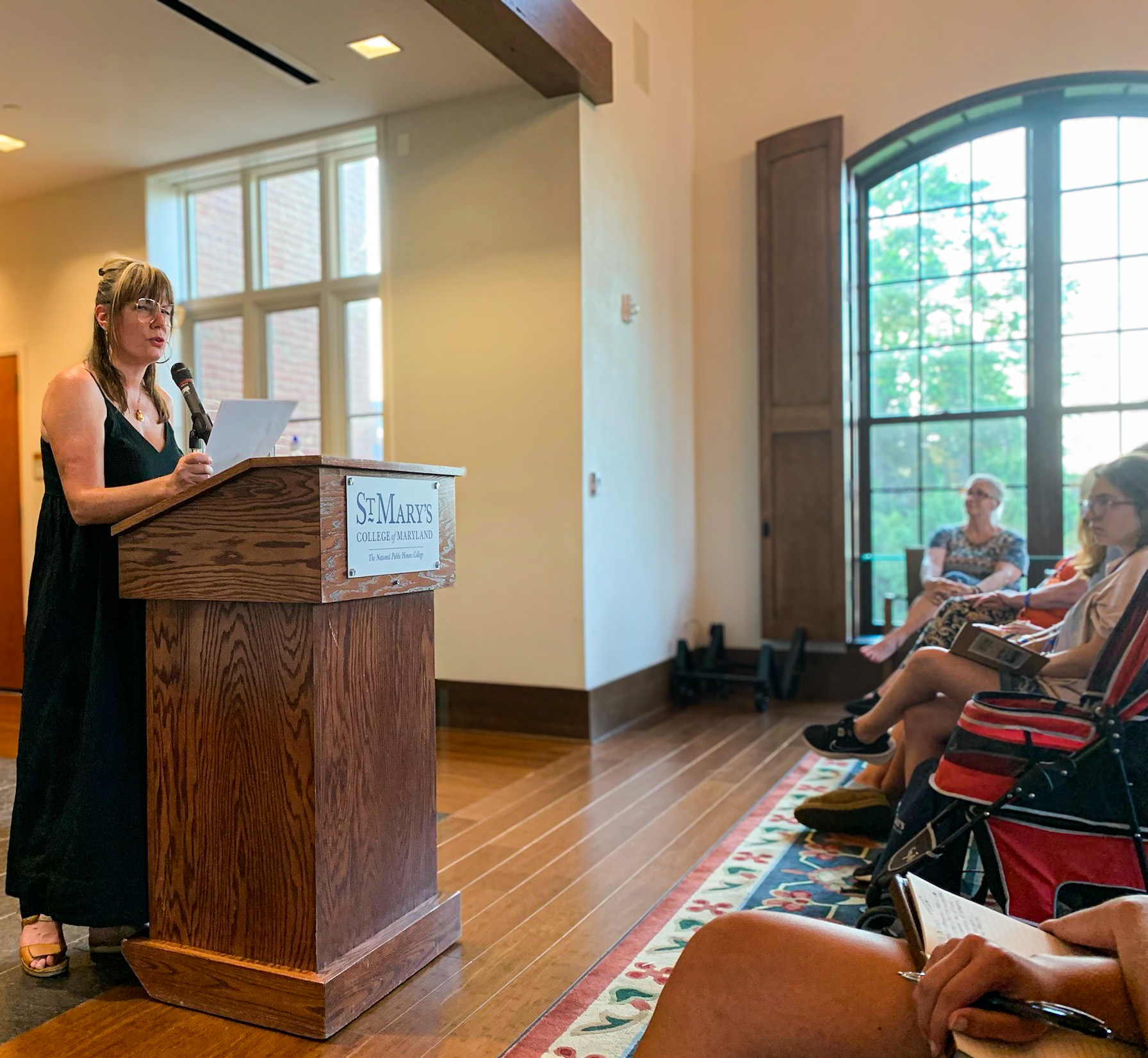 A woman stands at a podium labeled "St. Mary's College," reading from a paper to an audience seated in a sunlit room.