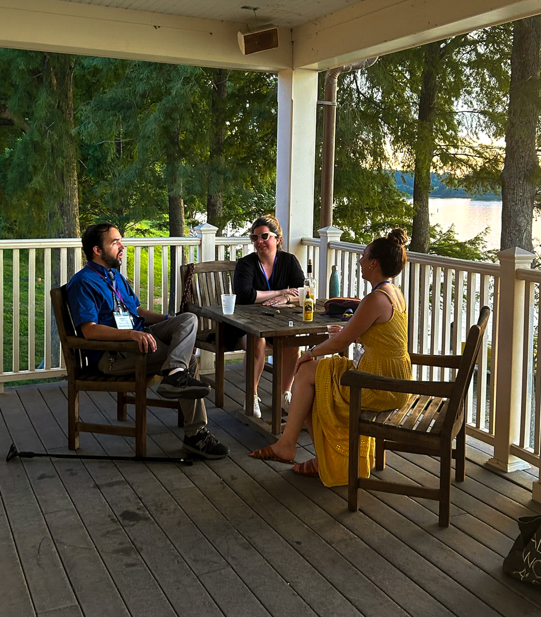 Three people sit and talk around a wooden table on a porch overlooking trees and a lake, with drinks on the table and sunlight in the background.