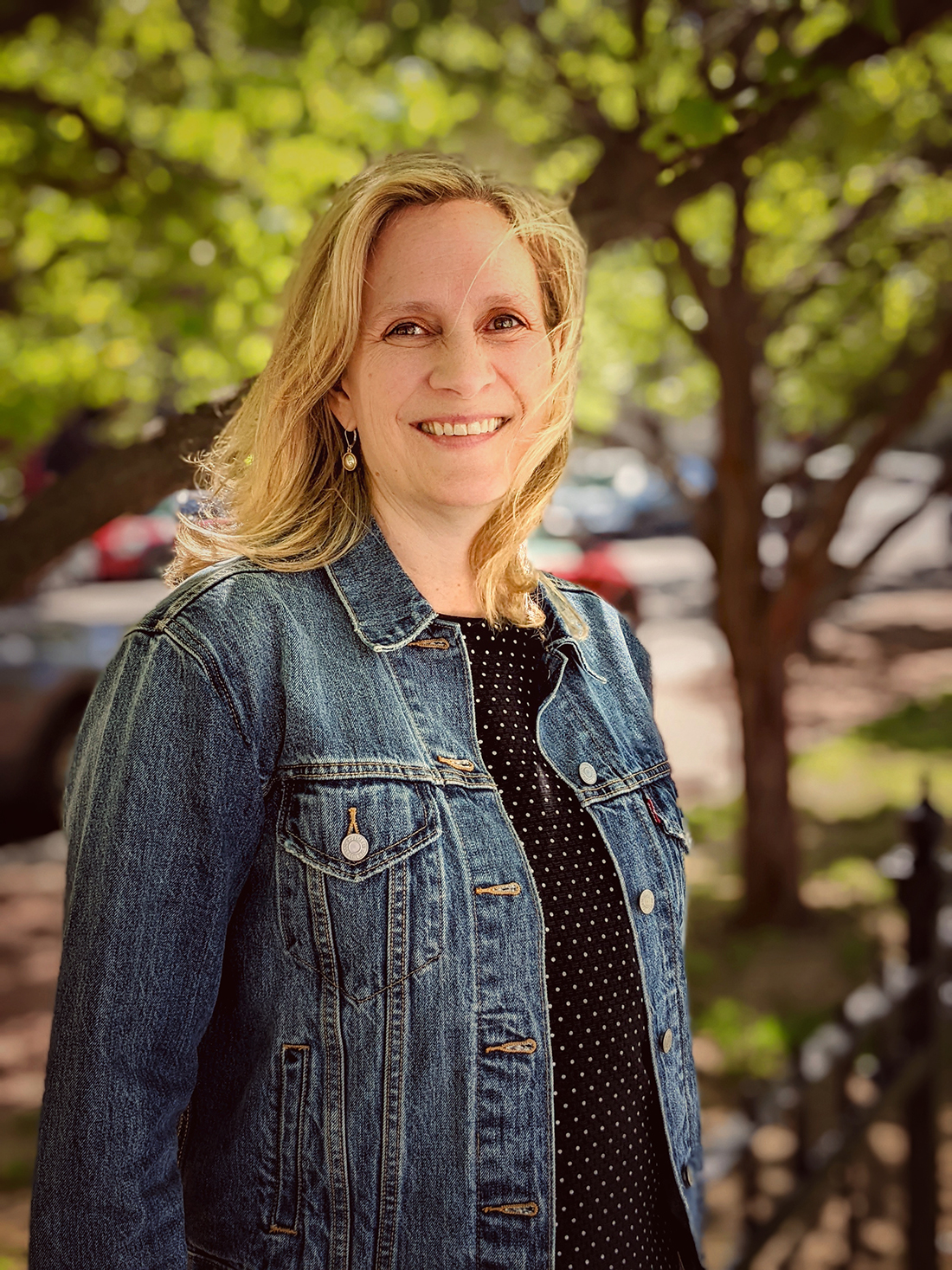 A woman with blonde hair wearing a denim jacket and black dress with white dots stands outdoors in front of leafy trees, smiling at the camera.