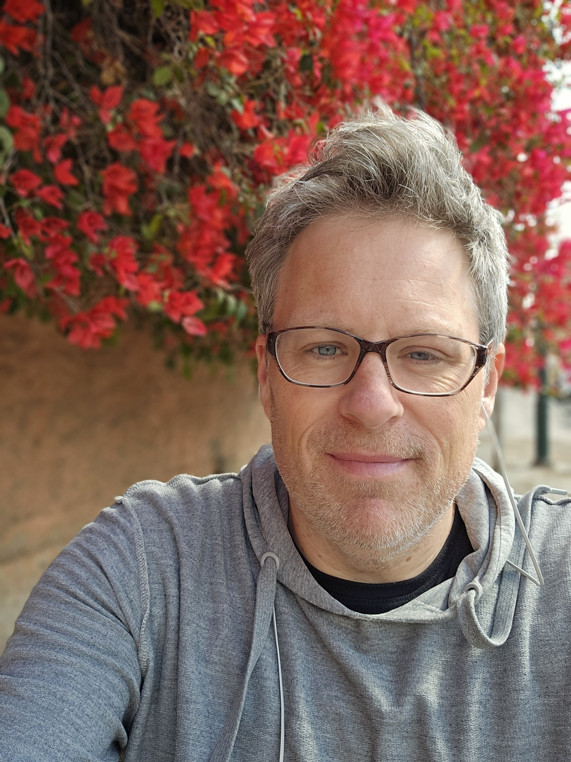 A man with gray hair and glasses wearing a gray hoodie stands in front of a wall covered with bright red flowers.
