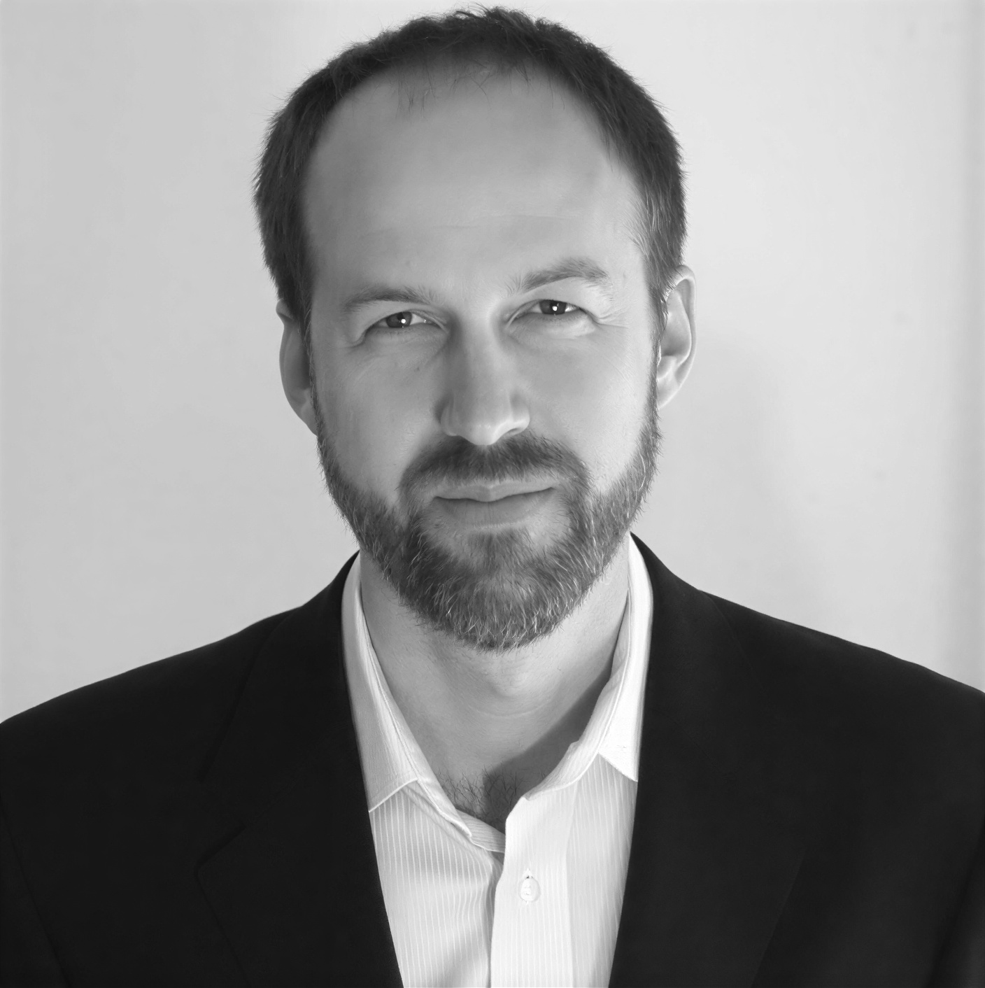 A man with short hair and a beard, wearing a dark blazer over a light-colored shirt, poses against a plain background.