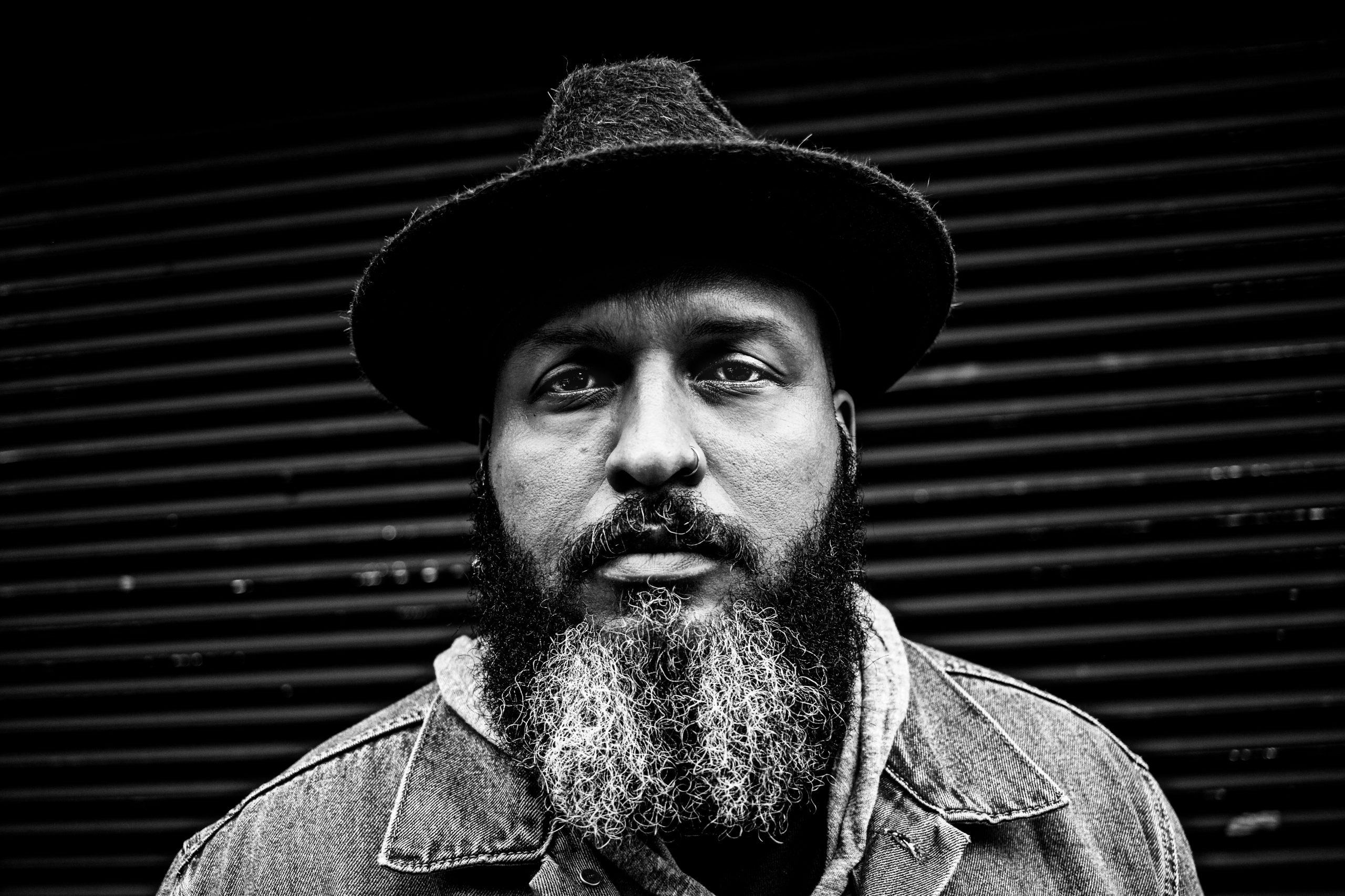 Black and white portrait of a bearded person wearing a hat and denim jacket, standing in front of a ribbed metal background. The image emphasizes texture and contrast, highlighting the person's contemplative expression.