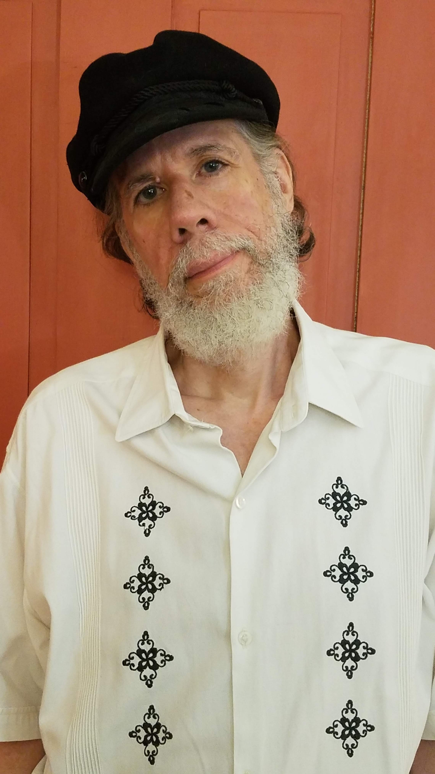 Elderly man with a white beard wearing a white embroidered shirt and a black cap stands in front of a red background.