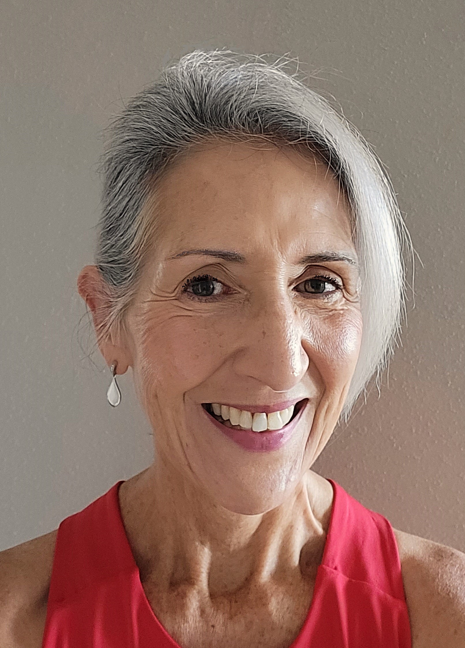 Smiling woman with short gray hair, wearing a red sleeveless top and small teardrop earrings, stands against a plain beige background.