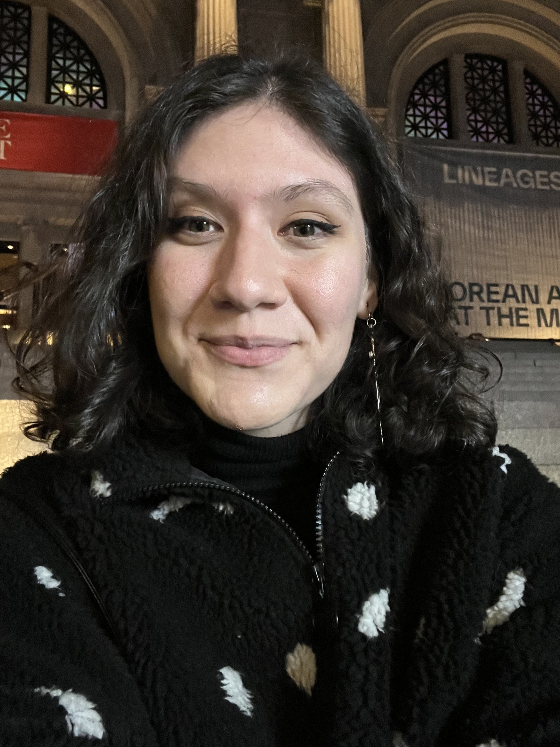 A person with curly hair and a black sweater with white spots smiles indoors, with columns and arched windows visible in the background.