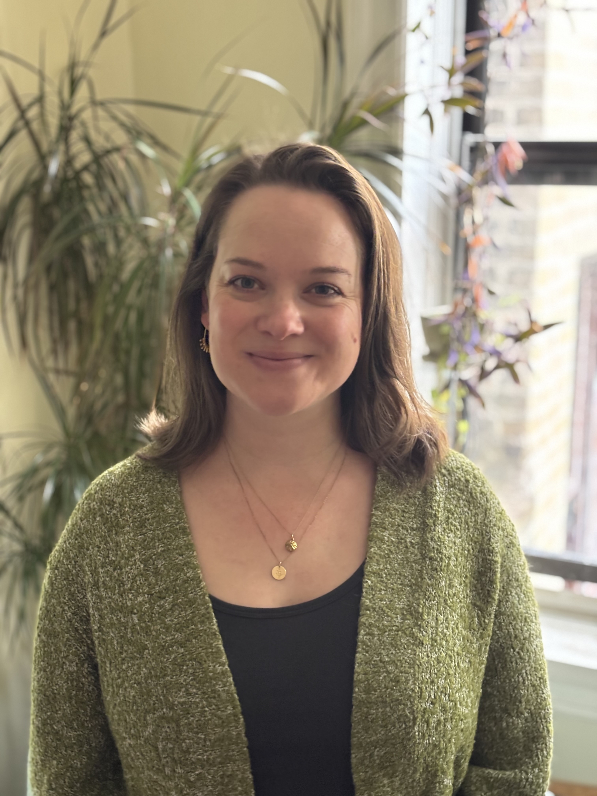 A woman with shoulder-length brown hair, wearing a green cardigan and gold necklaces, stands indoors in front of leafy plants and a sunlit window.