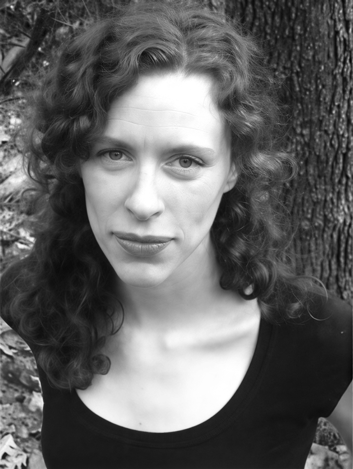 A woman with curly hair and a neutral expression stands outdoors in front of a tree, wearing a black top. The photo is in black and white.