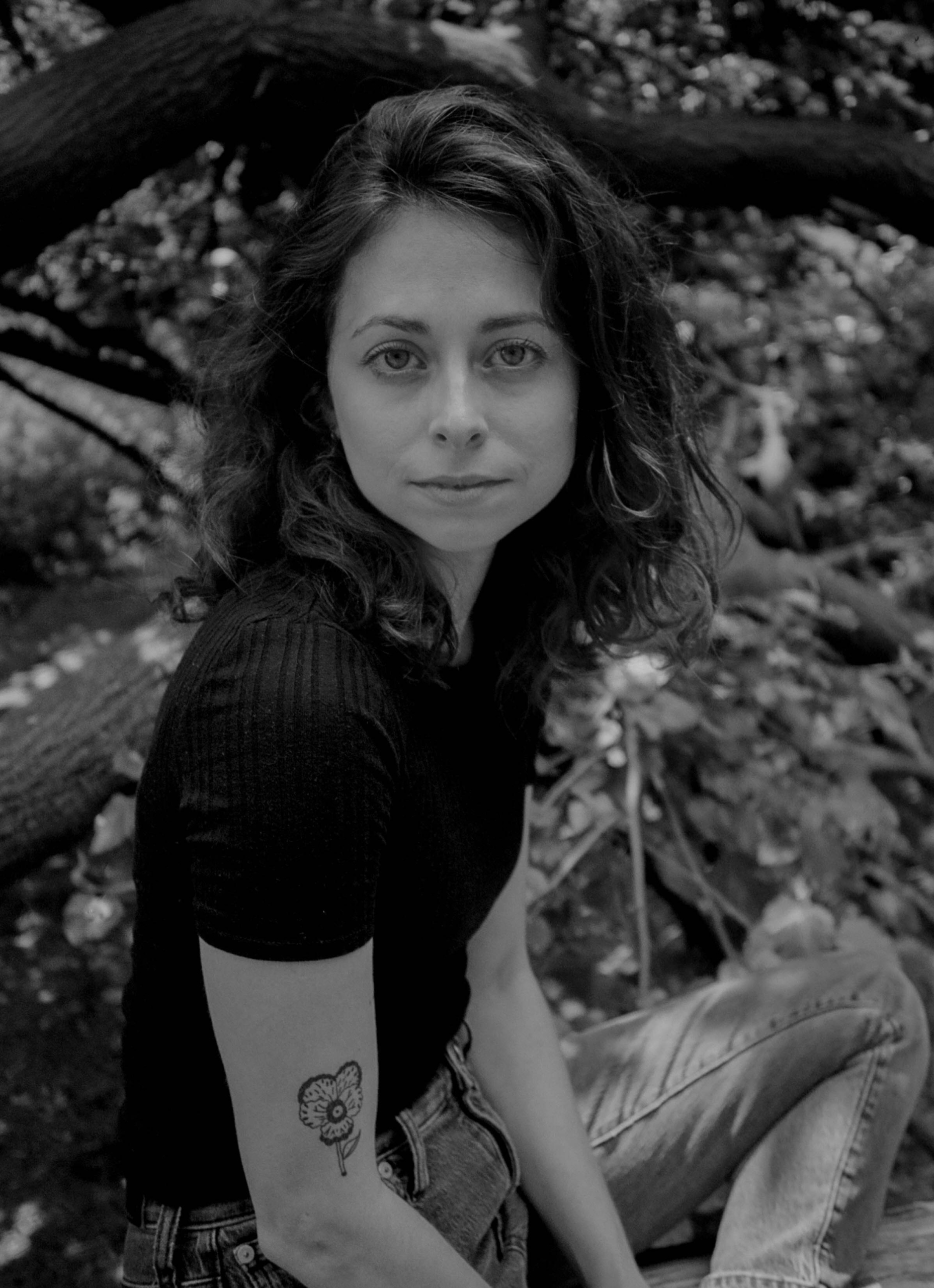 A woman with wavy hair and a flower tattoo on her arm sits outdoors in front of tree branches, looking at the camera. The image is in black and white.