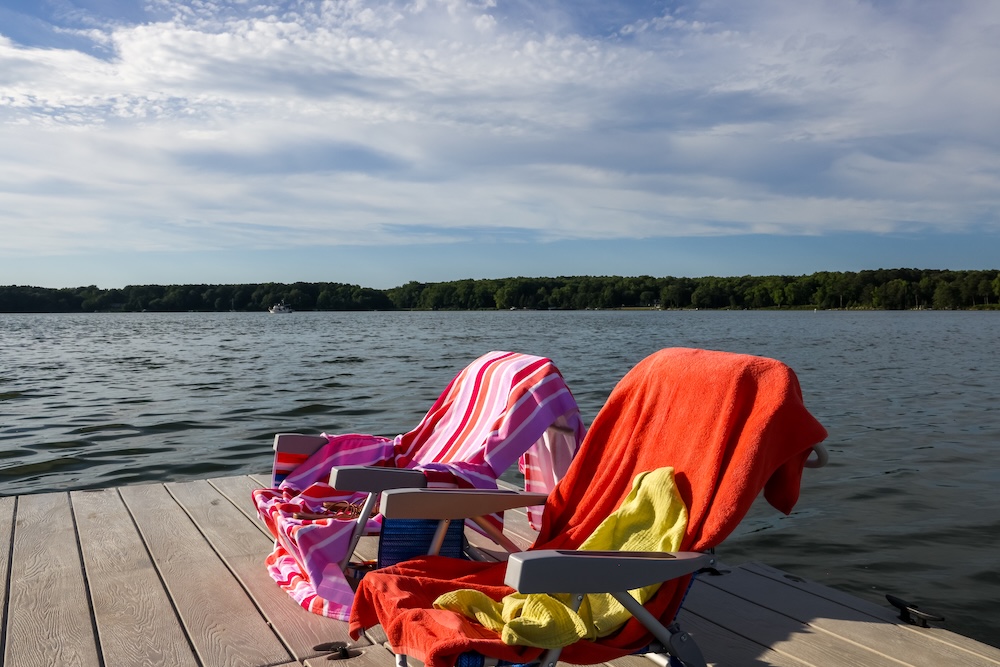 Two lounge chairs with colorful towels are set on a wooden dock by a lake, with water and trees in the background under a partly cloudy sky.