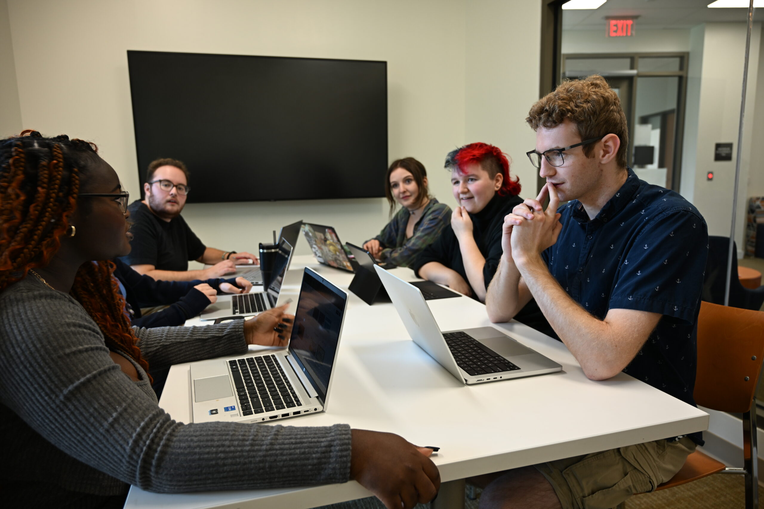 Five people sit around a table with laptops, engaged in discussion in a modern meeting room with a large screen on the wall.