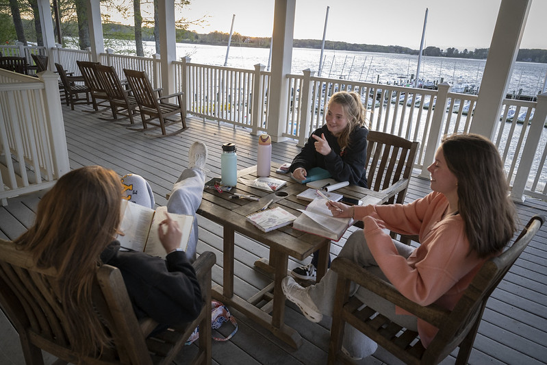 Three young women sit around a wooden table on a porch by the water, talking and studying with notebooks and drinks in front of them.