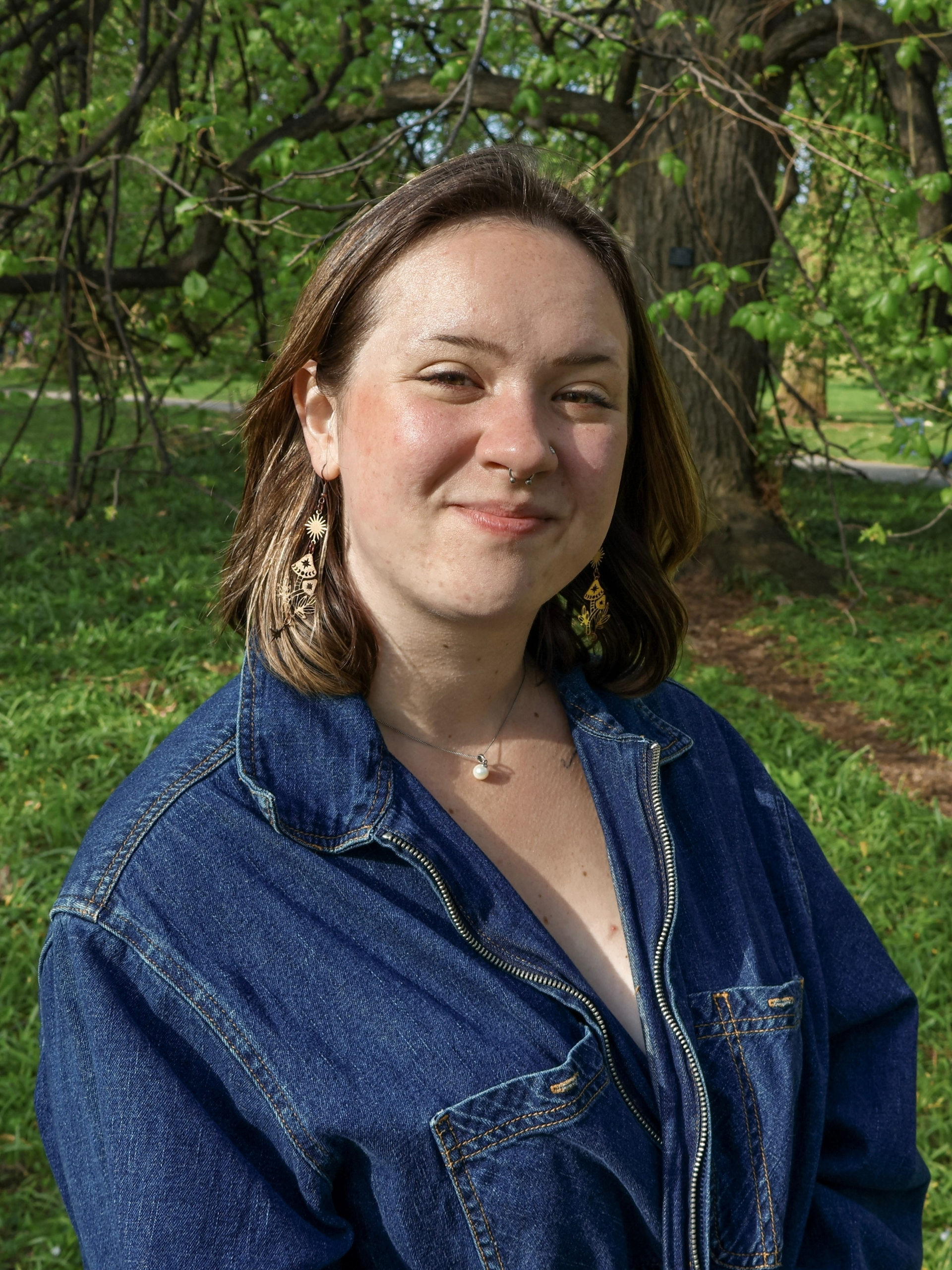 A person with short brown hair wearing a blue denim jacket and gold earrings stands outdoors in a green, wooded area.