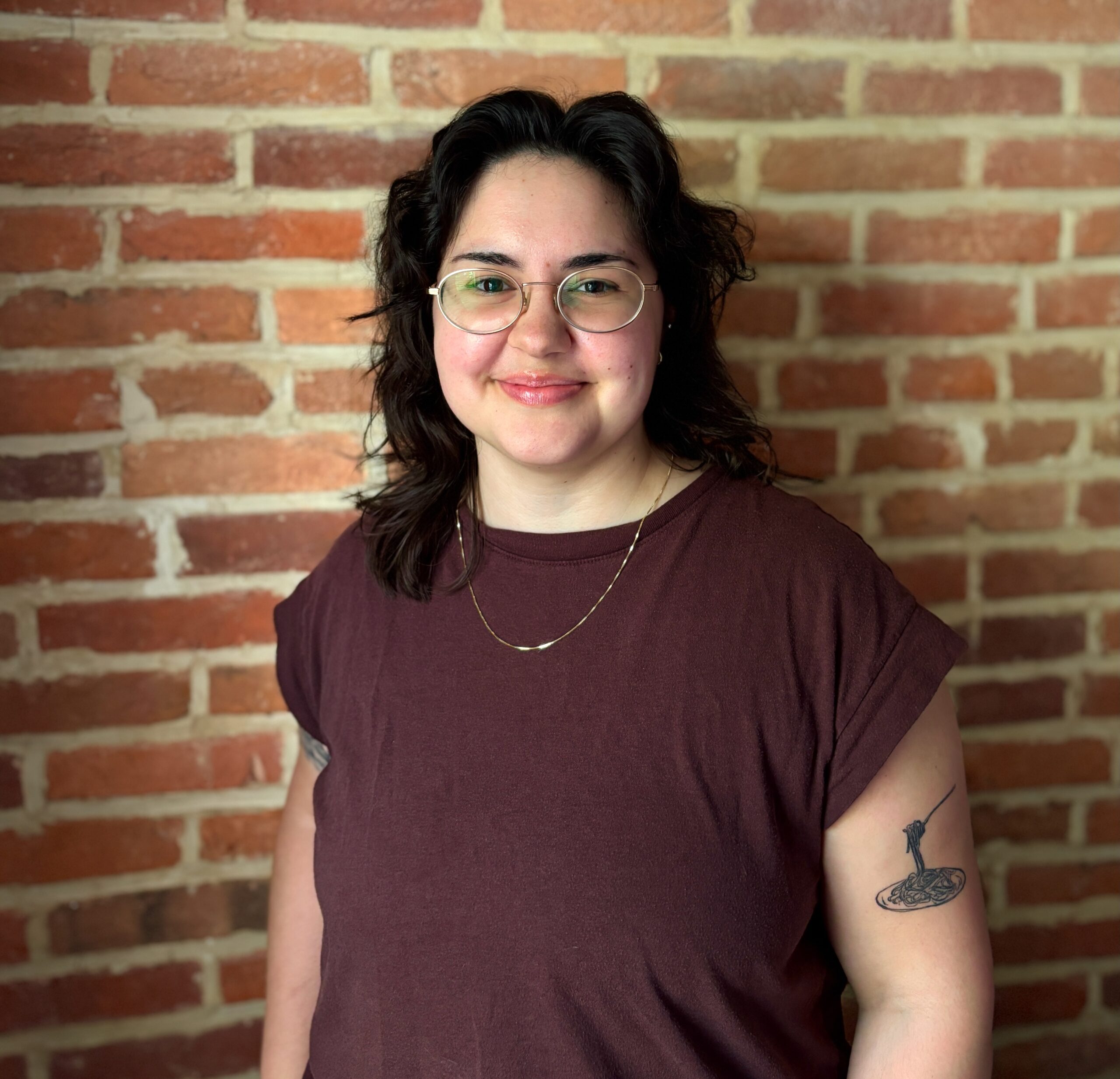 A person with medium-length dark hair, glasses, and a maroon shirt stands in front of a brick wall, smiling. They have a tattoo on their upper left arm and wear a thin necklace.