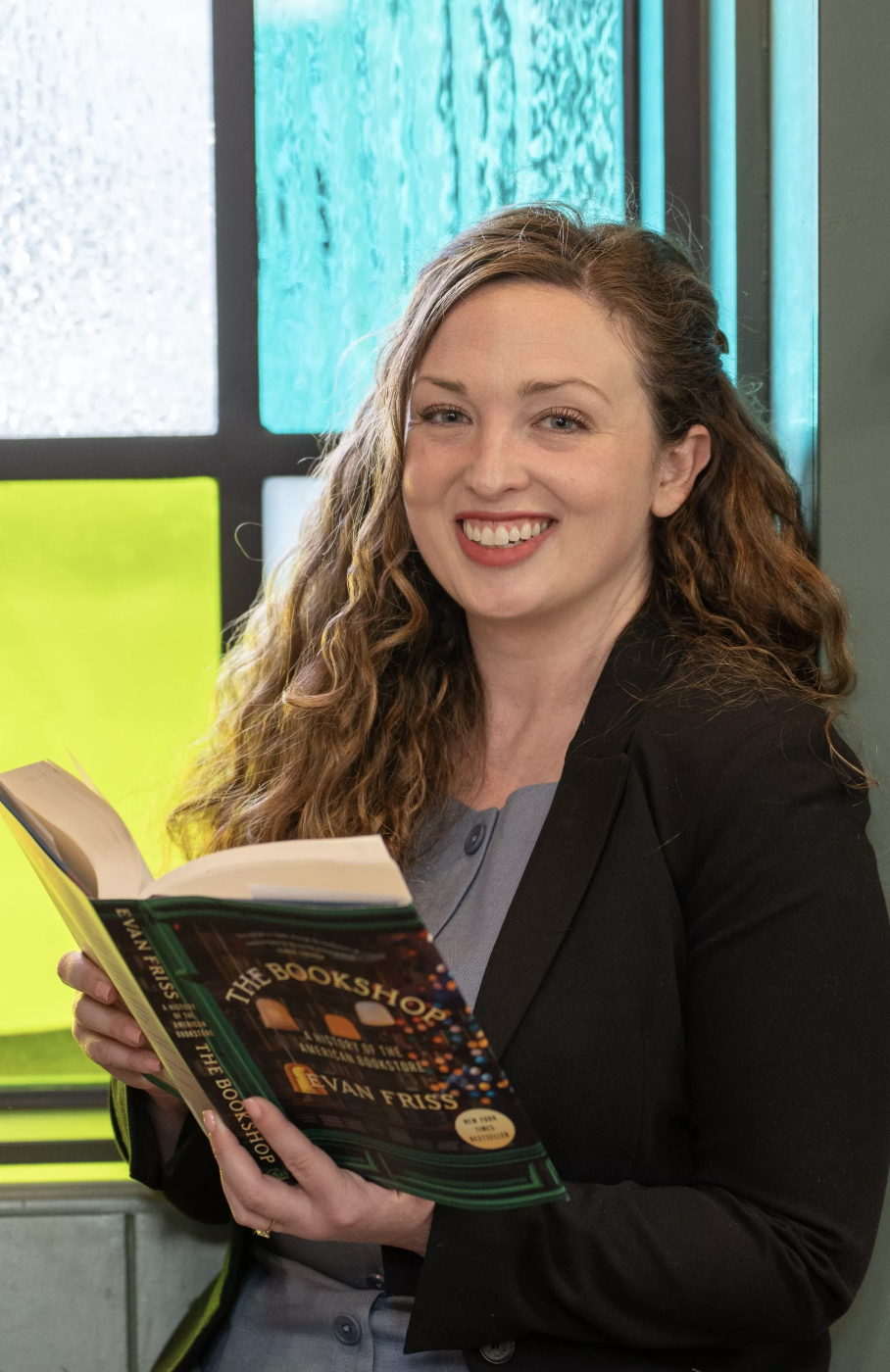 A woman with long curly hair, wearing a black blazer, is smiling while holding and reading "The Bookshop of Second Chances" by Jackie Fraser indoors near colorful windows.