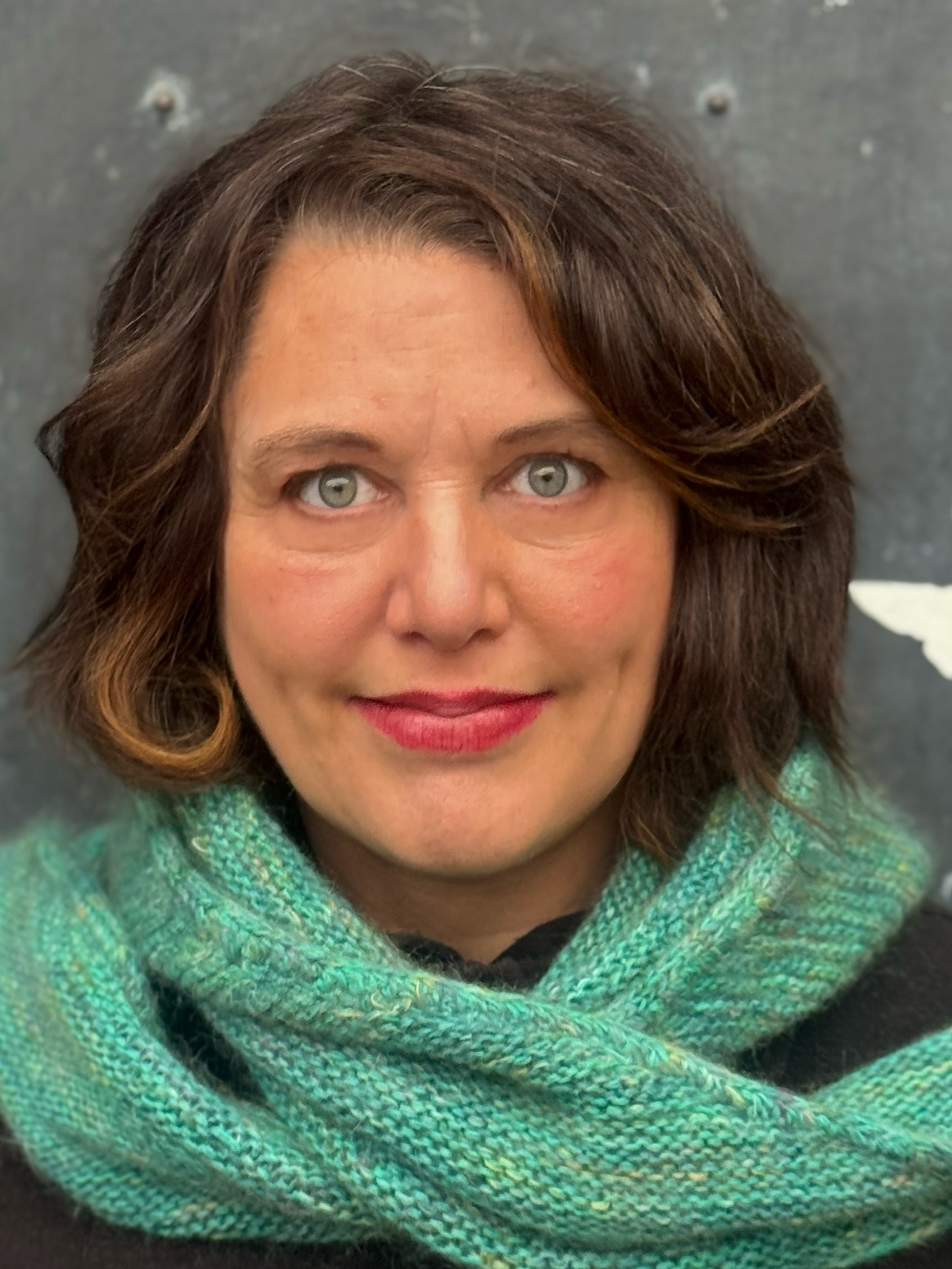 Woman with shoulder-length brown hair and green eyes wears a teal knit scarf and black top, looking directly at the camera with a neutral expression against a gray background.
