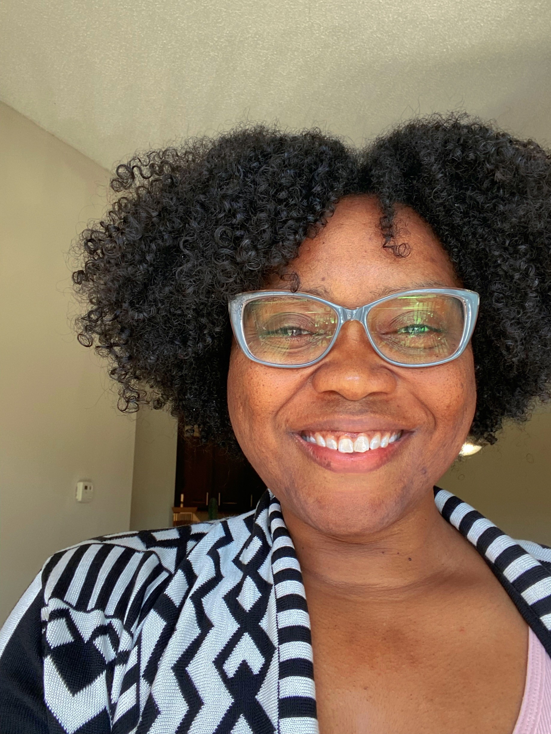A person with curly hair and glasses smiles at the camera, wearing a black and white patterned sweater.