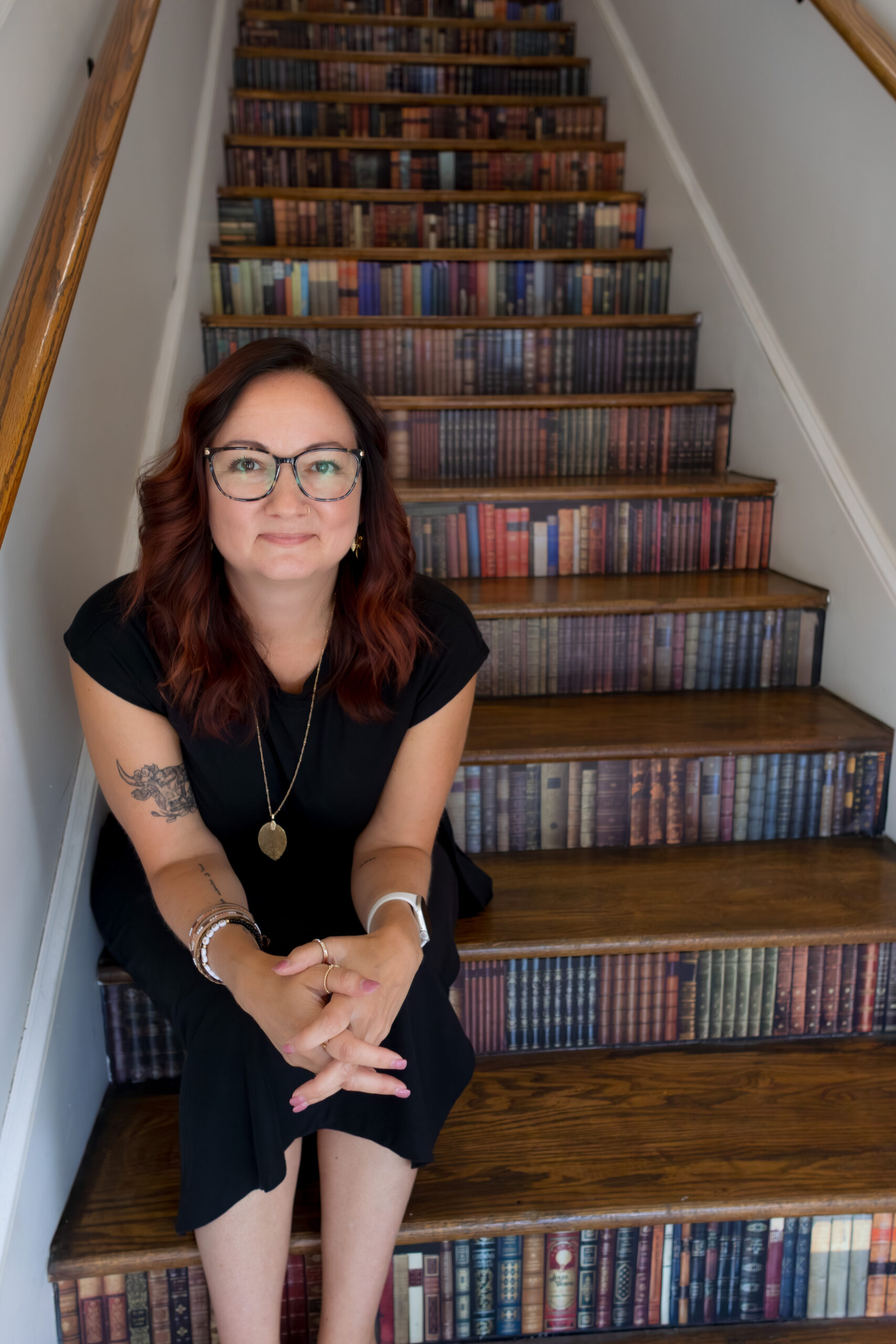 A woman with glasses and a black dress sits on wooden stairs decorated with images of colorful book spines.