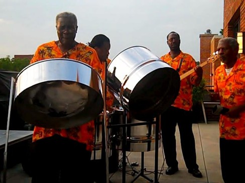 Four musicians wearing orange patterned shirts play steel drums and a bass guitar outdoors.