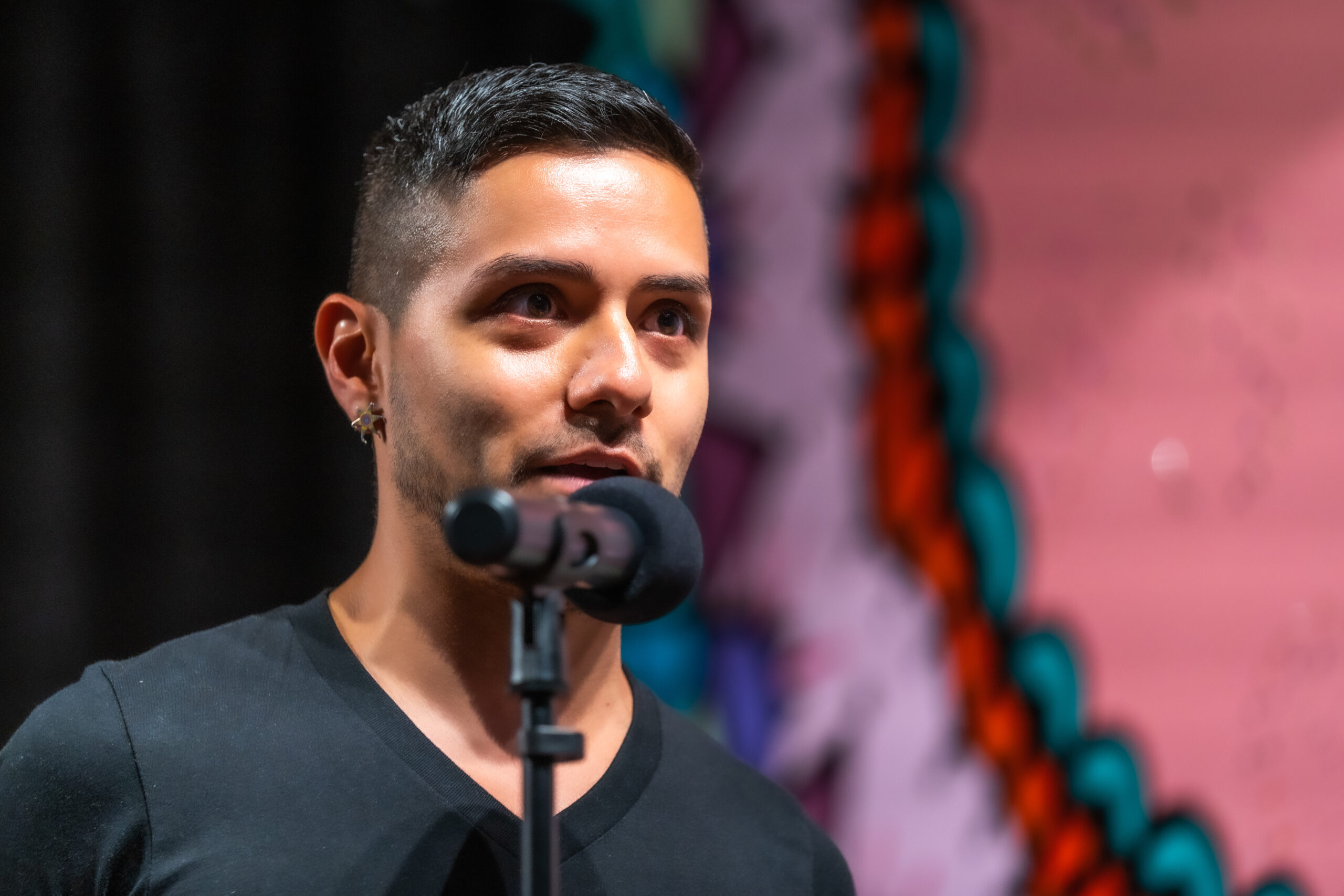 A man with short hair and a black shirt speaks into a microphone, standing in front of a colorful, abstract background.