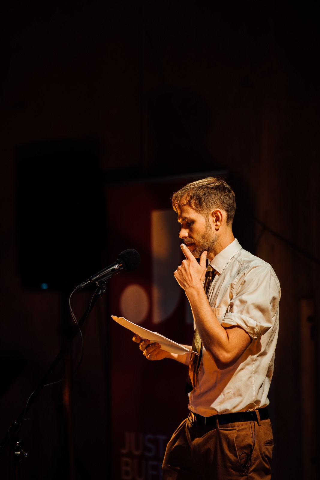A man in a white shirt and tie stands at a microphone, holding papers and touching his chin, appearing thoughtful on a dimly lit stage.