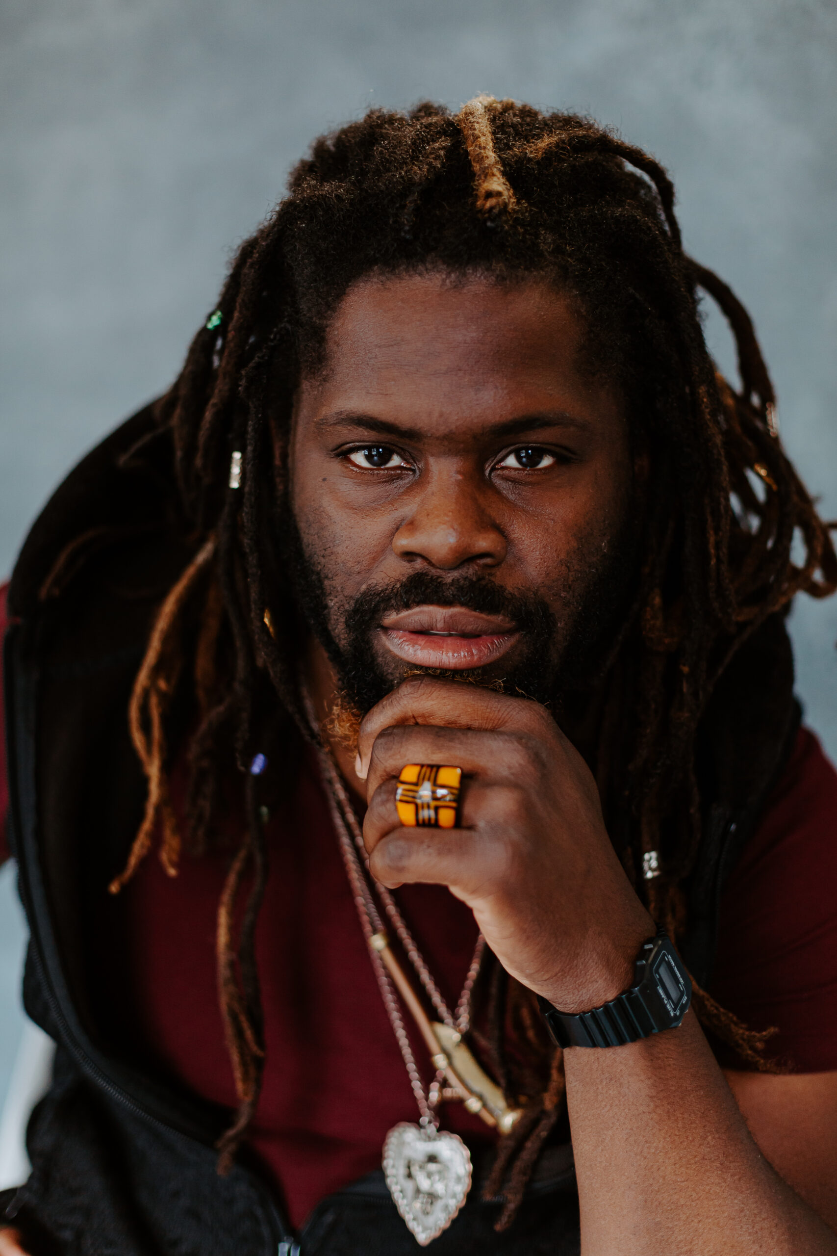 A man with long dreadlocks, wearing a maroon shirt, black vest, rings, necklaces, and a black watch, rests his chin on his hand while looking at the camera.