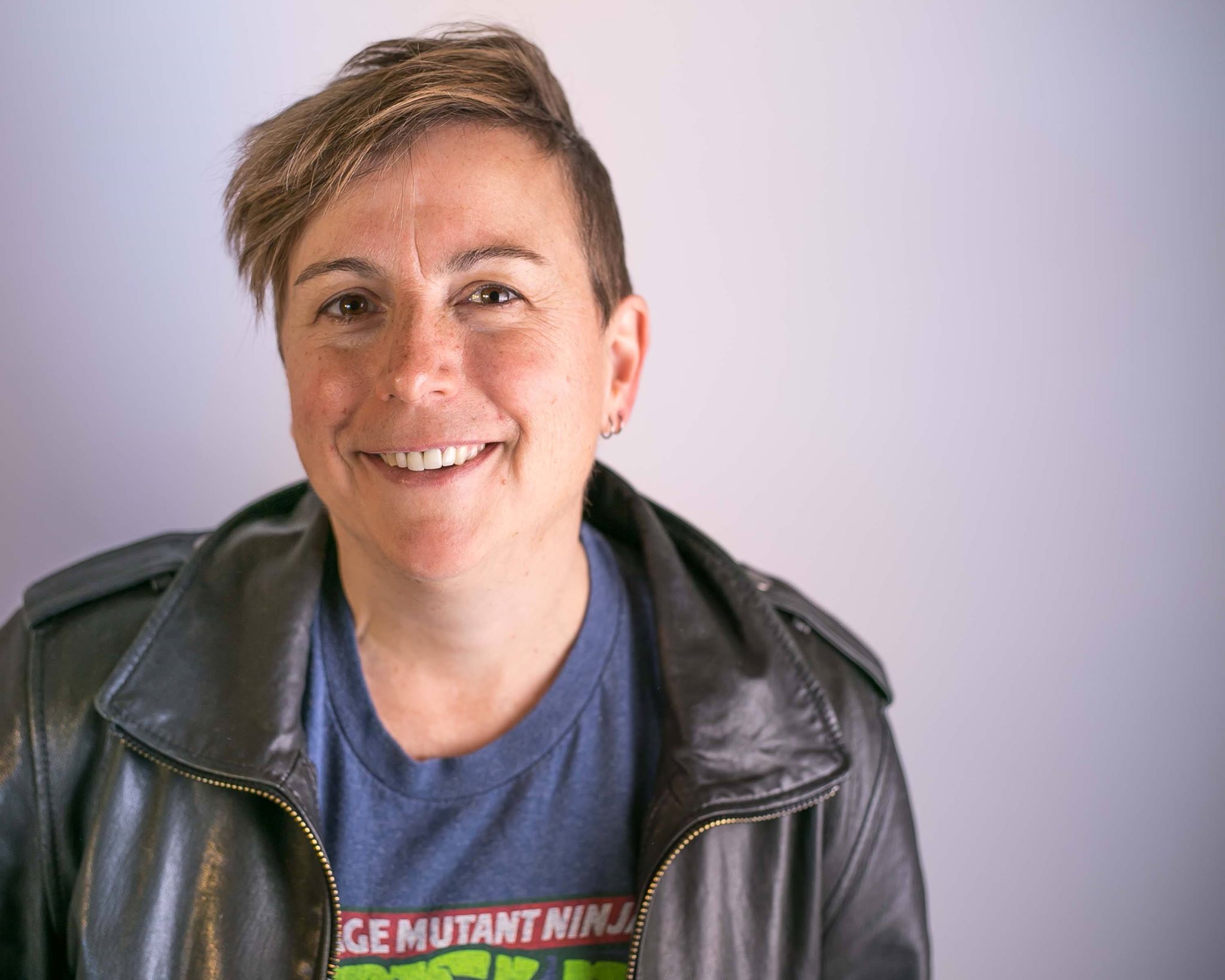Person with short brown hair, wearing a black leather jacket and a Teenage Mutant Ninja Turtles t-shirt, smiling in front of a plain light-colored background.