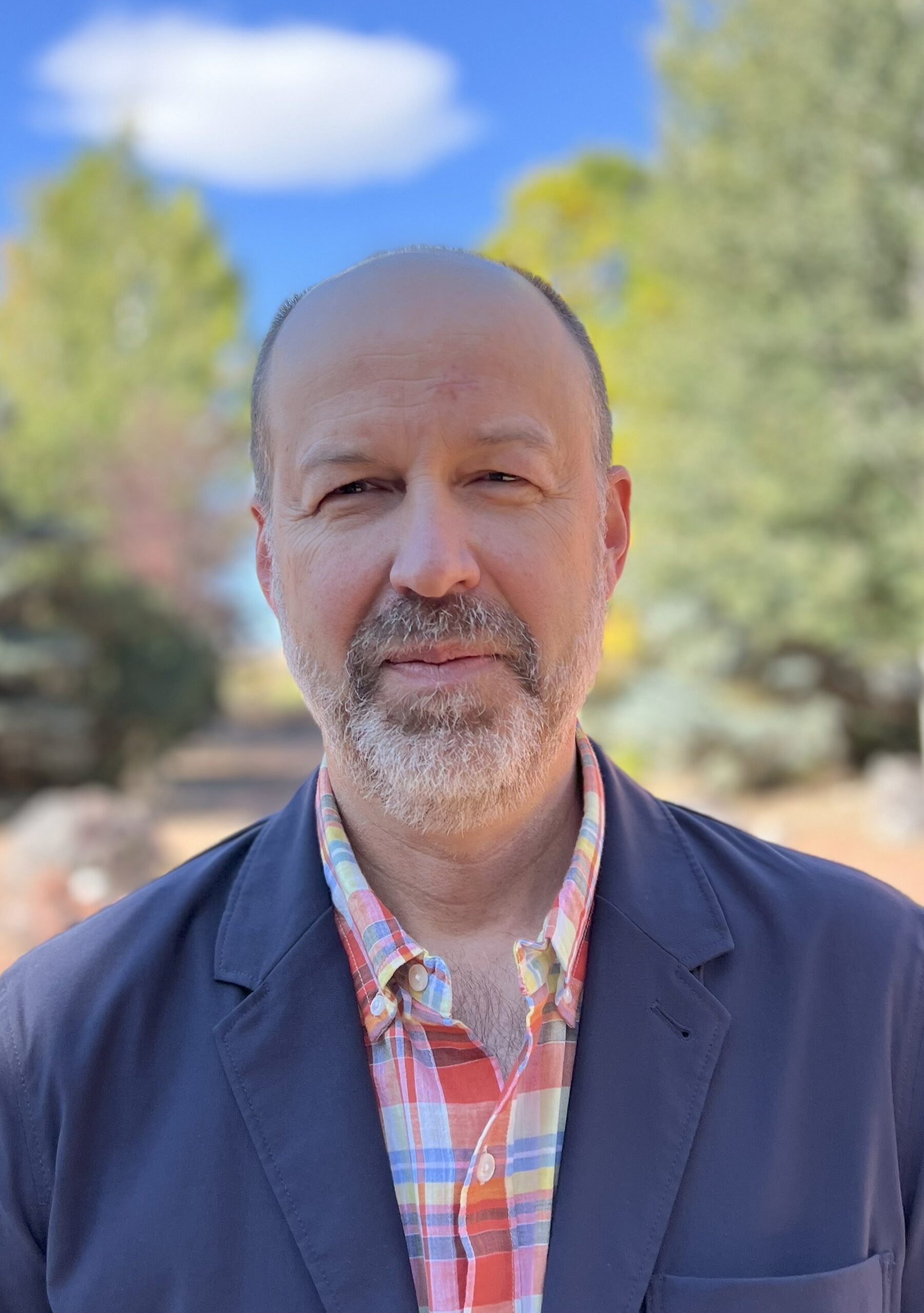 A middle-aged man with a beard stands outdoors, wearing a plaid shirt and navy jacket. Blurred trees and a blue sky are visible in the background.
