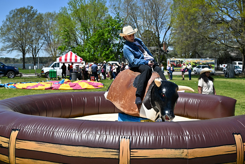 An SMCM student wearing a cowboy hat riding a mechanical bull at the World Carnival, with people, tents, and trees visible in the background.
