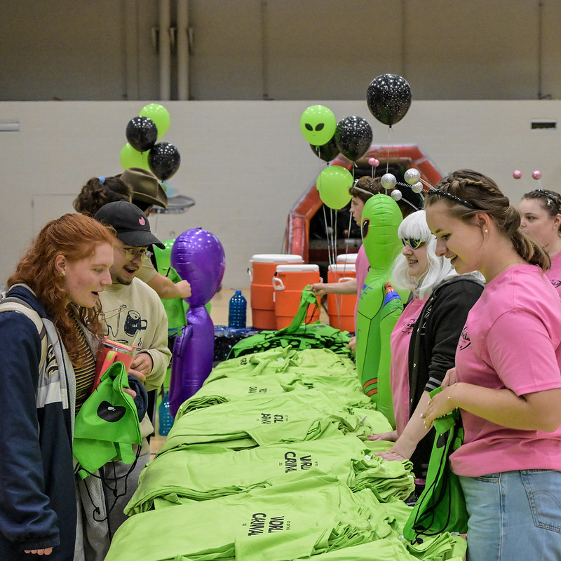 Students stand at a table covered with bright green t-shirts, with inflatable aliens and black-green balloons in the background at the World Carnival.