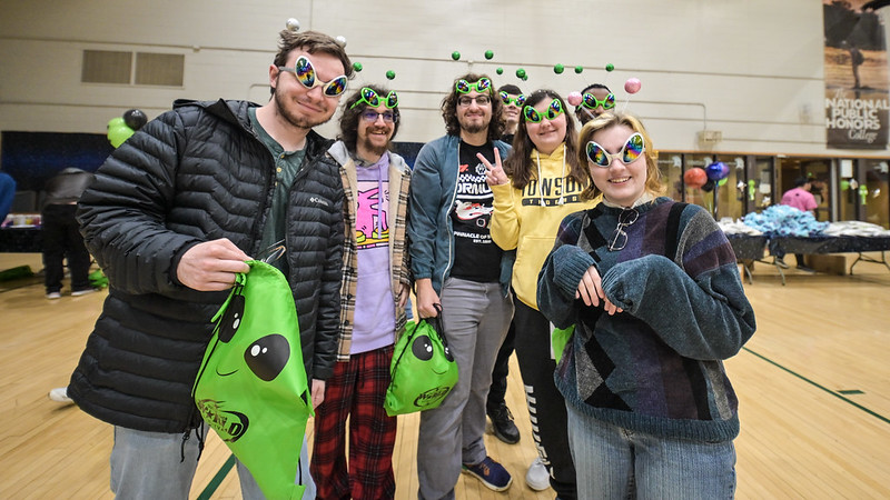 Five students wearing alien-themed headbands and glasses stand together in a gymnasium, holding green bags with cartoon alien faces at the World Carnival.