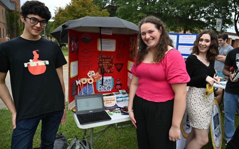Three students stand by a table with a display board and laptop the Club Involvement Fair, with trees and other booths in the background.