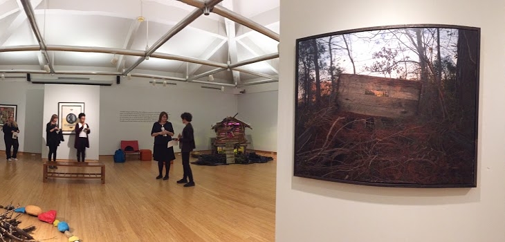 17_boydenGallery A gallery with people standing and observing art; a large photograph of a wooded scene with a wooden structure is displayed on the right wall.