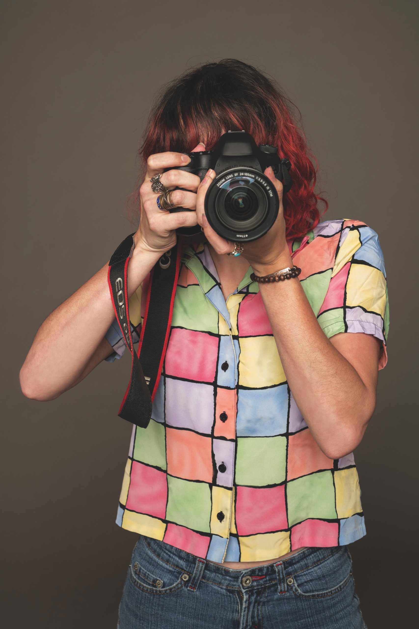 2131-1344-adj Person wearing a colorful checkered shirt and blue jeans holding a camera up to their face, standing against a neutral background.