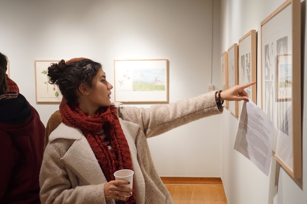 A person wearing a red scarf and beige coat holds a cup and points at framed artwork on a gallery wall, holding a sheet of paper in the other hand.