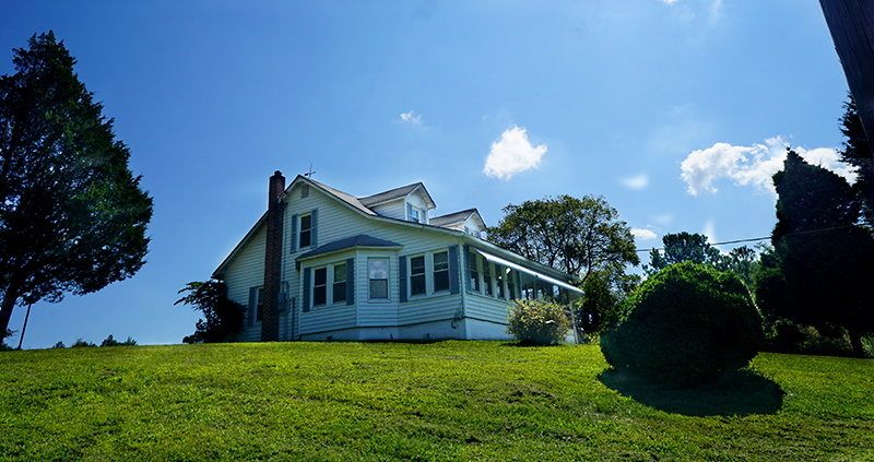 A white two-story house with a porch sits on a grassy hill under a clear blue sky with a few clouds and surrounding trees.