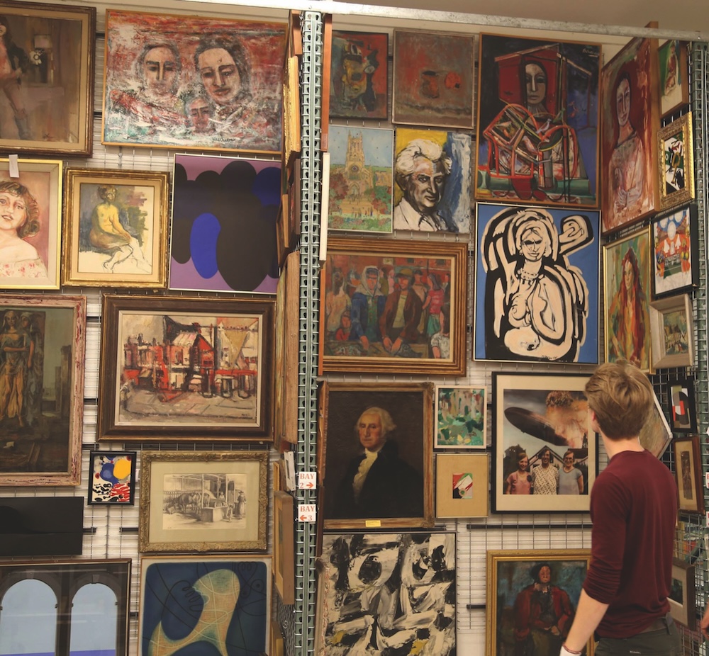 A person stands in front of a wall filled with various framed paintings and artworks displayed in a gallery or storage setting.