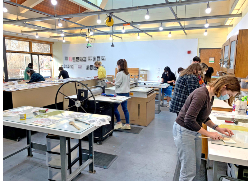 MH-129-Printmaking-BookArts-2-scaled People work on art projects at various tables in a well-lit studio, using tools and materials; artwork and photos are displayed on the walls.