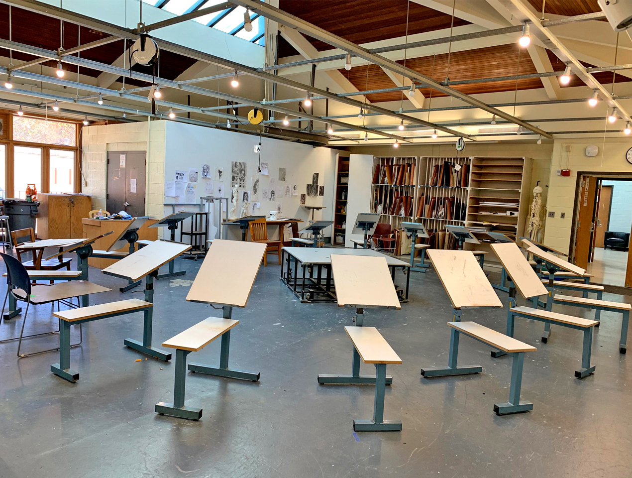 MH-135_Drawing-2 An art classroom with several tilted drawing tables, stools, scattered chairs, and art supplies; sketches and books line the shelves and walls in the brightly lit studio space.