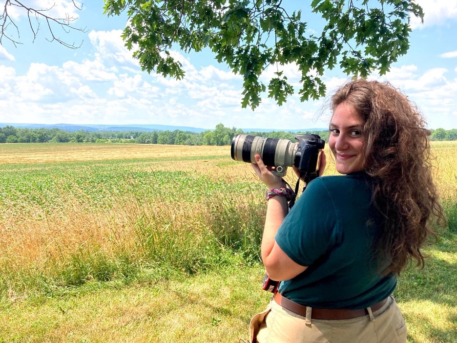 A person with long curly hair holds a DSLR camera with a telephoto lens, standing in a grassy field under a tree on a sunny day.
