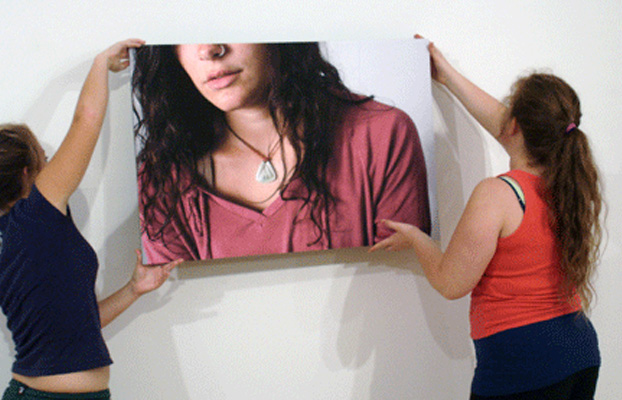Two women hang a large photograph on a white wall. The image shows a woman's chest and face partially out of frame, wearing a pink shirt and a pendant necklace.