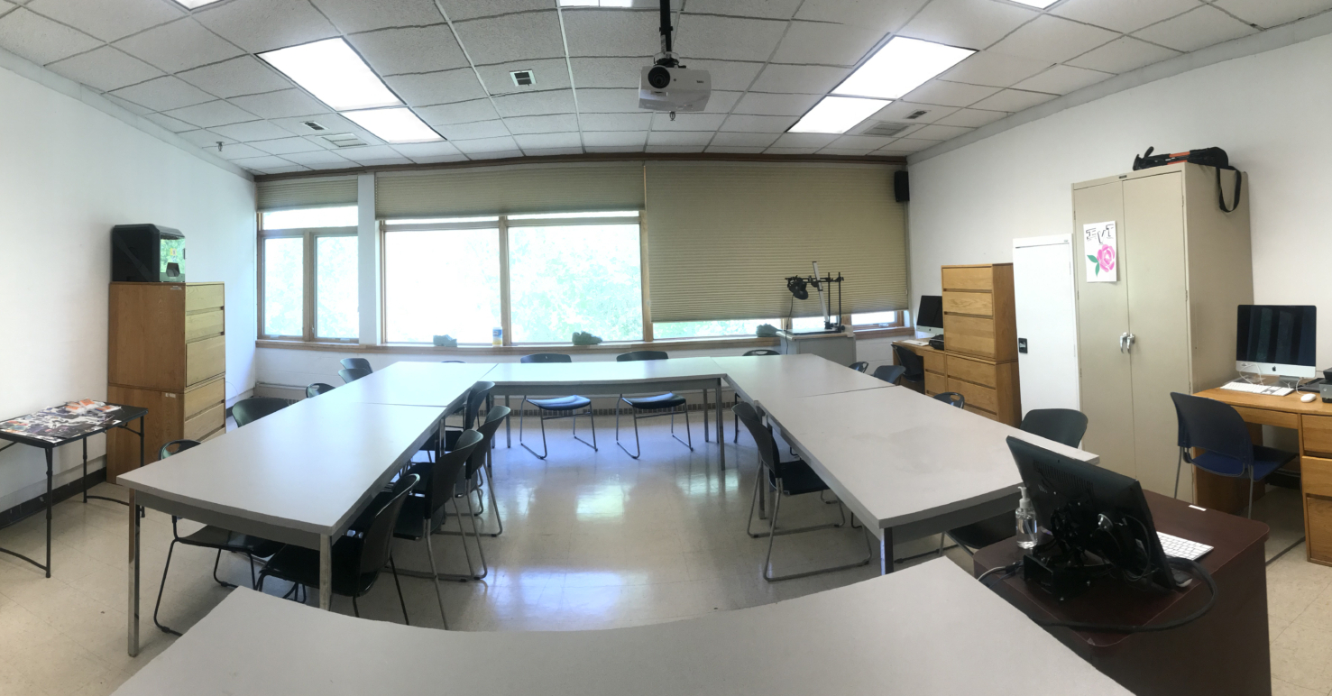 best105-1-scaled A classroom with U-shaped tables, chairs, cabinets, a computer, and large windows letting in natural light.