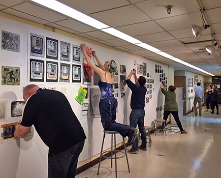 hallwallsCombo Several people hang and arrange artwork and prints on a long white wall in a hallway, preparing for an art display or exhibition.