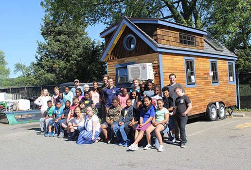 interdisciplinaryCombo A group of people, including children and adults, pose together in front of a small wooden house on wheels in an outdoor setting.