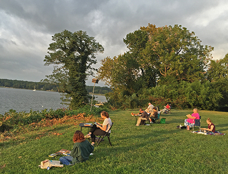 outdoorClassroomCombo Several people sit on a grassy hill, sketching or painting while facing a lake bordered by trees under a cloudy sky.