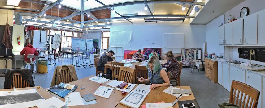 A group of people work on art projects at tables in a spacious, well-lit studio with art supplies and finished paintings visible around the room.