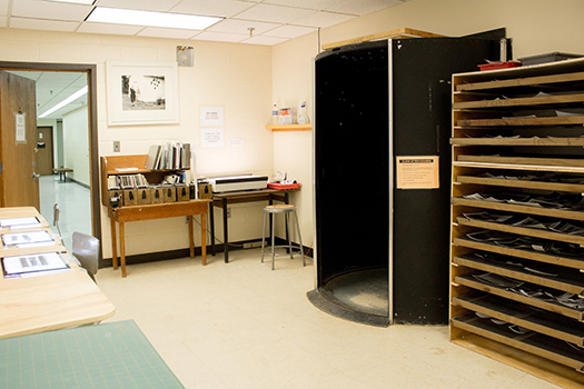 photoComboSmall02 An art classroom with a drafting table, shelves holding plastic trays, a darkroom booth, and a desk with art supplies against the wall.